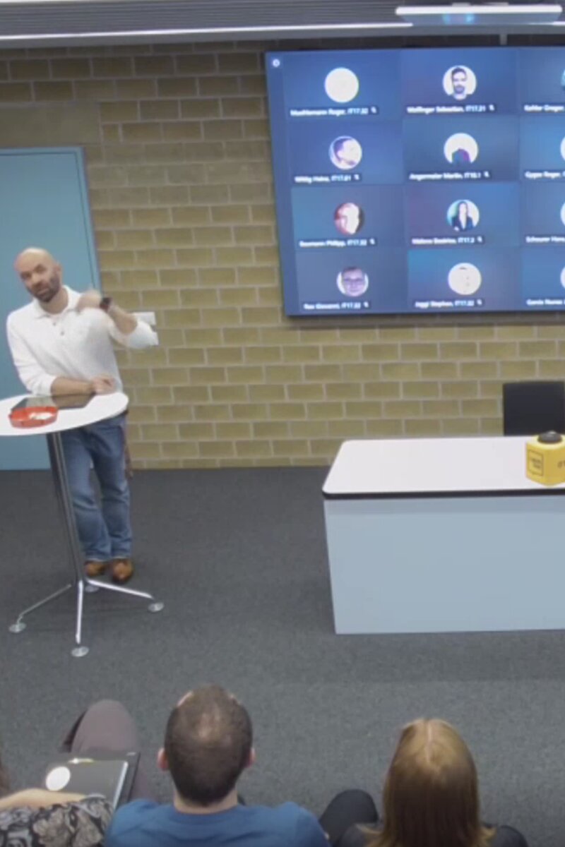 A presenter beside a video-call screen with seated attendees